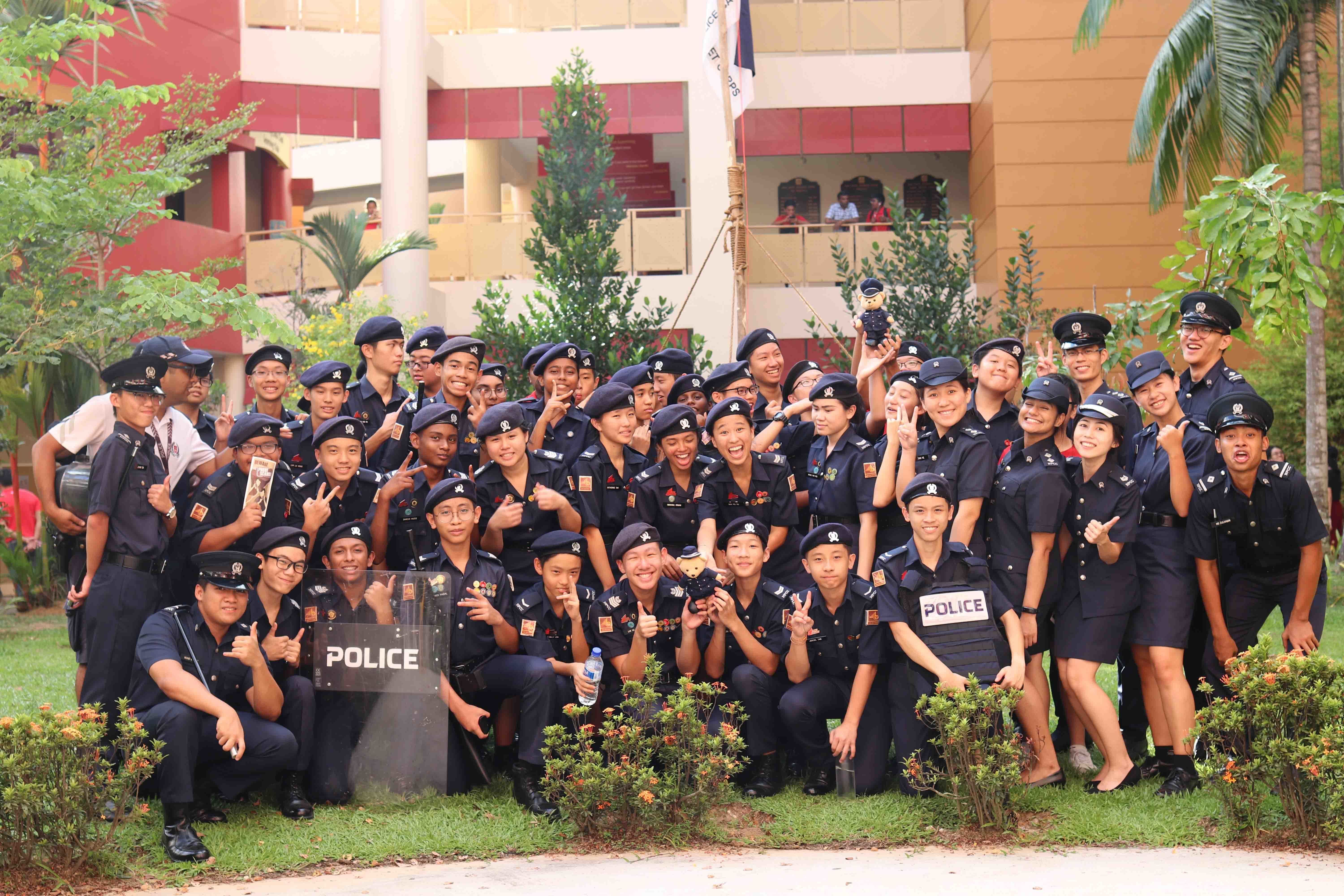 Large group of young people in dark navy police cadet uniforms posing outdoors near a red building.